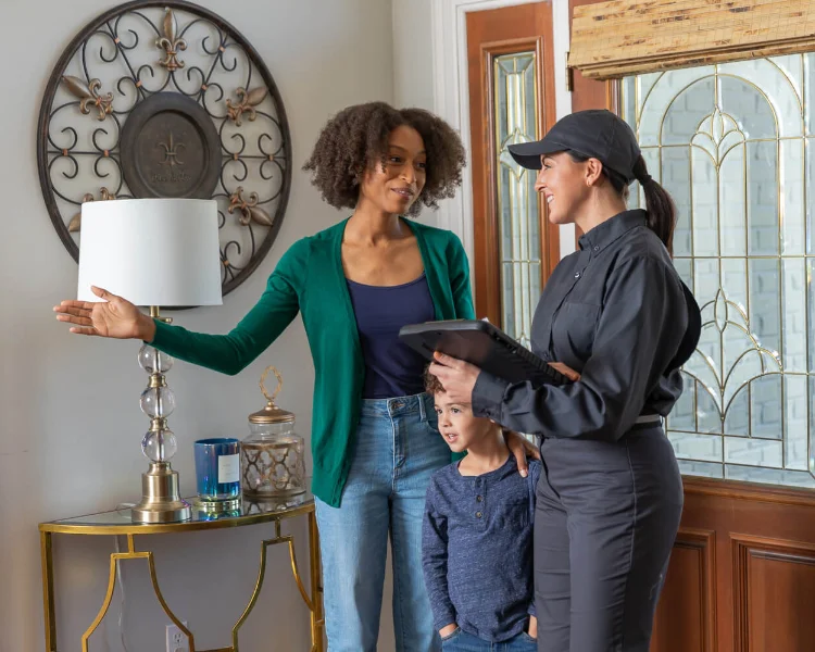 A female pest control technician holding a tablet while speaking with a homeowner and her young child near an entryway.